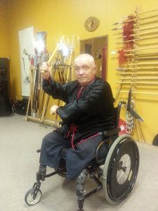 A student in a wheelchair training with a staff weapon before the weapons wall