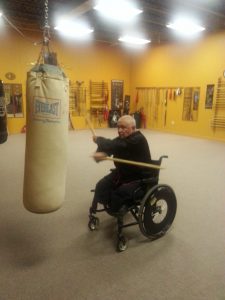 A student in a wheelchair working the heavy bag with a staff weapon
