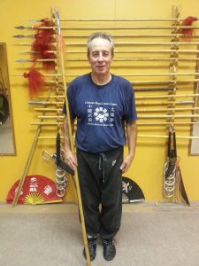 Student standing with a long spear before the weapons rack