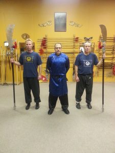 Three students in traditional dress holding glaive weapons before the weapons wall