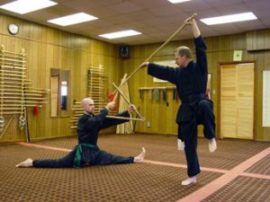 Two students in an acrobatic weapons drill, one in a full split, one airborne with a pole weapon raised