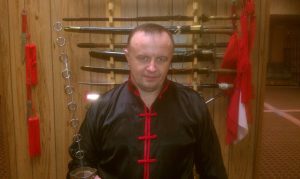 Student standing before the weapons rack, red tassels hanging beside curved blades