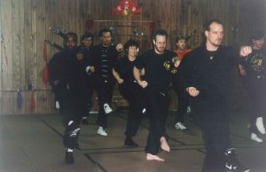 A row of students in black uniforms practicing kicking drills together in a wood-paneled training hall